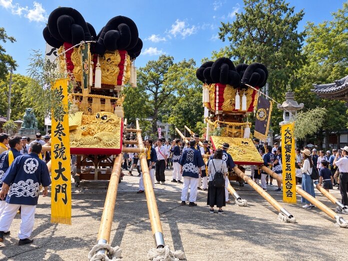 神社の境内に並ぶ2台の太鼓台。金色の龍の刺繍と黒い房飾りの付いた山車を、揃いの法被を着た担ぎ手たちが囲んでいる。